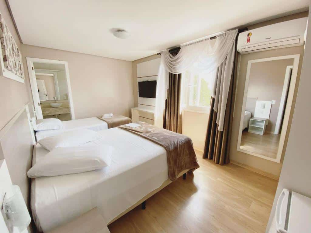 Quarto da Pousada Vovô Nino com uma cama de casal e uma de solteiro no canto esquerdo, e um espelho vertical no canto direito. Ao fundo é possível ver uma porta aberta com acesso ao banheiro, para representar pousadas em Gramado.