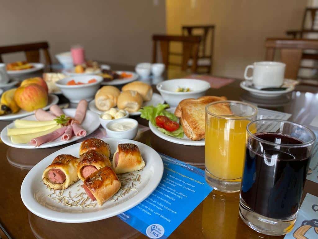 Mesa de café da manhã da Pousada Vovô Nino com diversos pratos dispostos. Há salsichas enroladas e assadas, mistos quentes, pães de sal, frutas, bolos, xícaras de café brancas e copos transparentes com sucos coloridos.