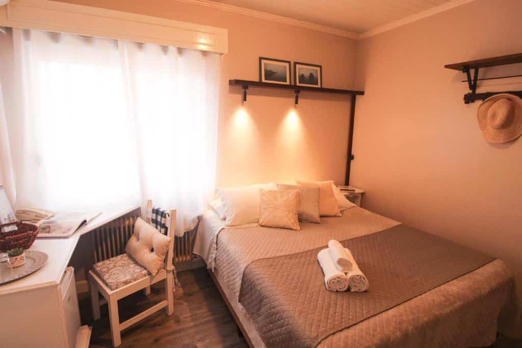 Quarto da Pousada Betânia com uma cama de casal à direita e uma mesa de canto branco com uma cadeira estofada à esquerda, para representar pousadas em Gramado.
