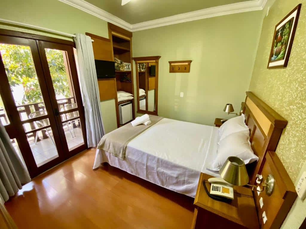 Quarto da Pousada Gramado com uma cama de casal à direita e portas duplas com acesso à varanda à esquerda. No local há também um frigobar, uma televisão e um espelho vertical na parede do fundo.