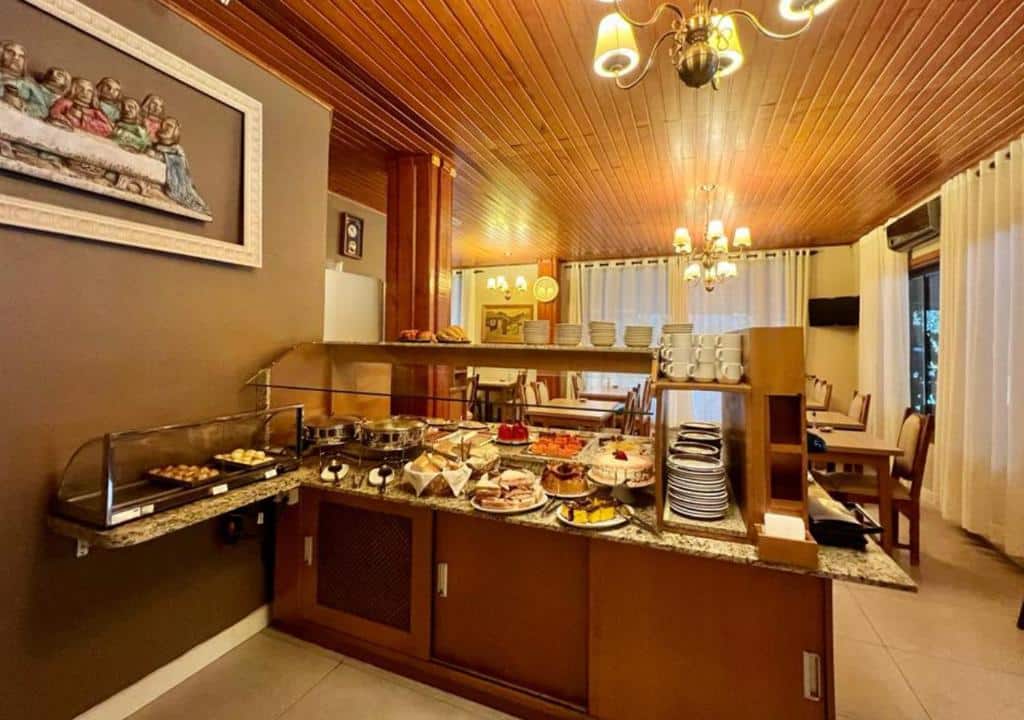 Refeitório da Pousada Gramado com um balcão repleto de alimentos no centro da imagem, Já bolos, pães e salgados, além de pratos e xícaras brancas empilhadas.