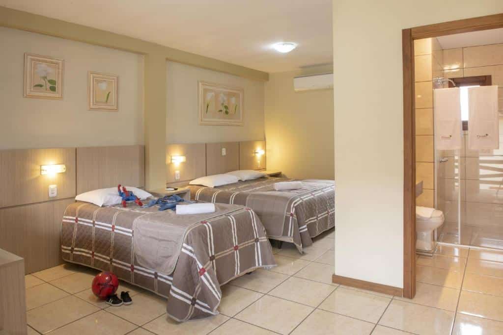 Suíte da Pousada Floratta Nossa Casa com uma cama de casal e uma cama de solteiro, onde um boneco do homem aranha está jogado em cima do travesseiro, e no chão há uma bola vermelha com dois tênis pretos pequenos. No lado direito da imagem há uma porta aberta que dá acesso ao banheiro com box, para representar pousadas em Gramado.