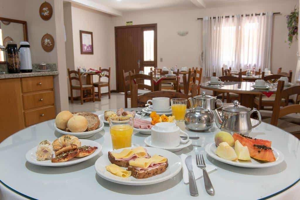 Refeitório da Pousada Floratta Nossa Casa com mesas redondas de madeiras e cadeiras ao redor. Bem ao centro há uma mesa arrumada para o café da manhã com diversos pratos: pães, frios, croissants, pães de queijo, frutas, sucos e café.
