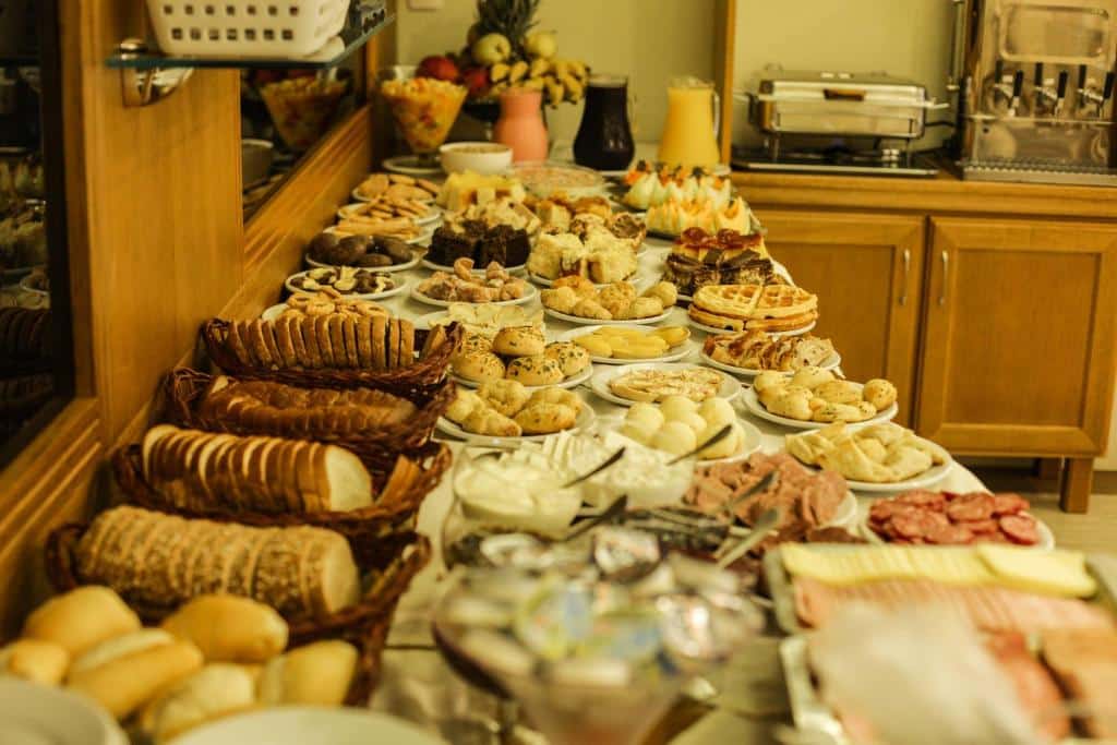 Uma mesa de bufê com vários pães, bolos, queijos, frios e frutas expostos em uma área de jantar bem iluminada, servido na Pousada das Papoulas, para representar pousadas em Gramado.