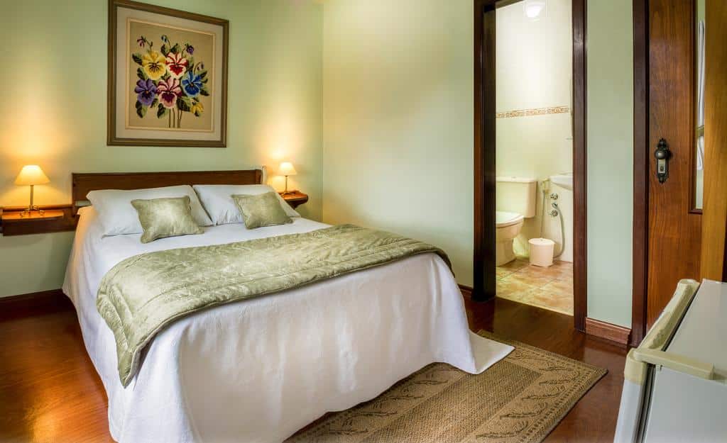 Suíte da Pousada Mikolay com uma cama de casal à esquerda e uma porta com acesso ao banheiro no lado direito, sobre a cama há uma quadro de flores coloridas, para representar pousadas em Gramado.