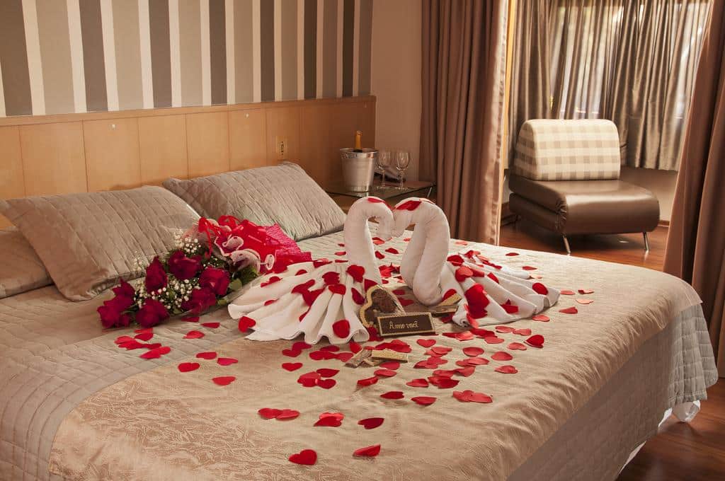 Cama de casal da Pousada Bernardete com pétalas de rosa espalhadas sobre a cama, além de um buquê deitado, juntamente com dois origamis de toalhas brancas em formato de cisnes de frente um para o outro, para representar pousadas em Gramado.