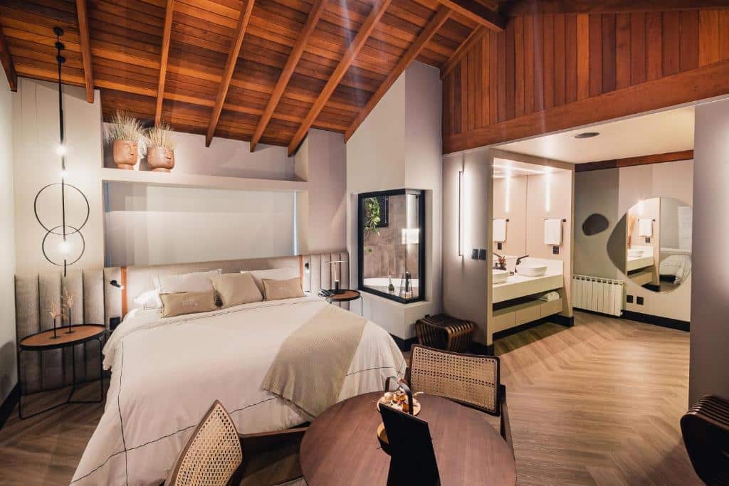 Suíte espaçosa da Pousada Bella Terra com uma cama de casal à esquerda, uma mesa redonda de madeira com duas cadeiras à frente da cama, e no lado direito, duas pias brancas com acesso ao banheiro que possui uma janela de vidro, de onde é possível ver a banheira, para representar pousadas em Gramado.