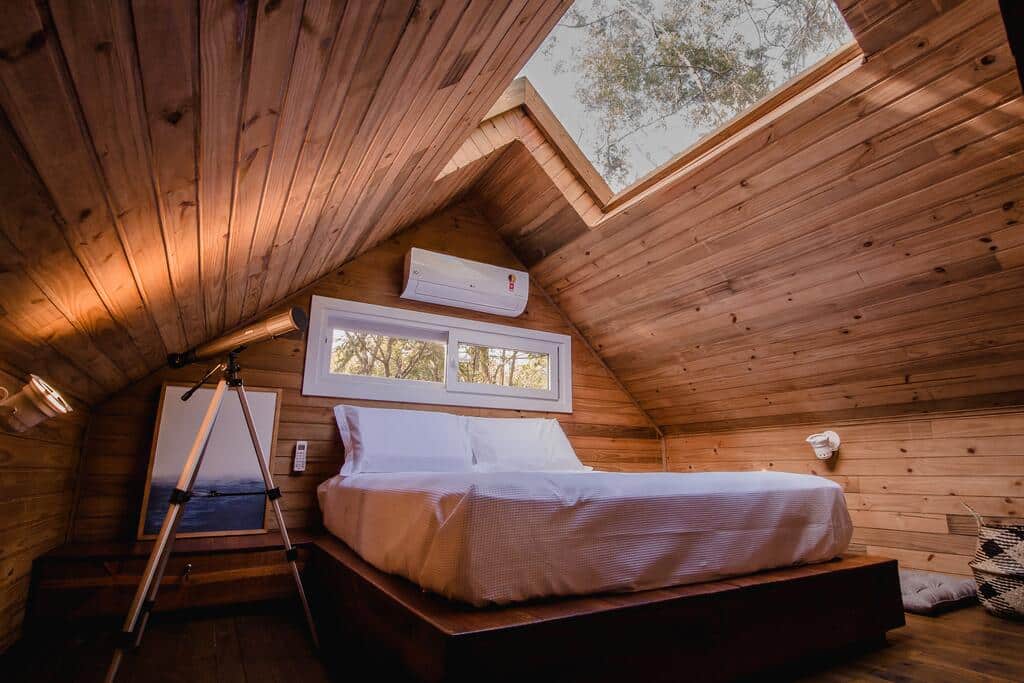 Suíte da Vila Nambu com uma cama de casal no centro do quarto, um telescópio ao lado e um teto solar com vista para as árvores, em Gramado.