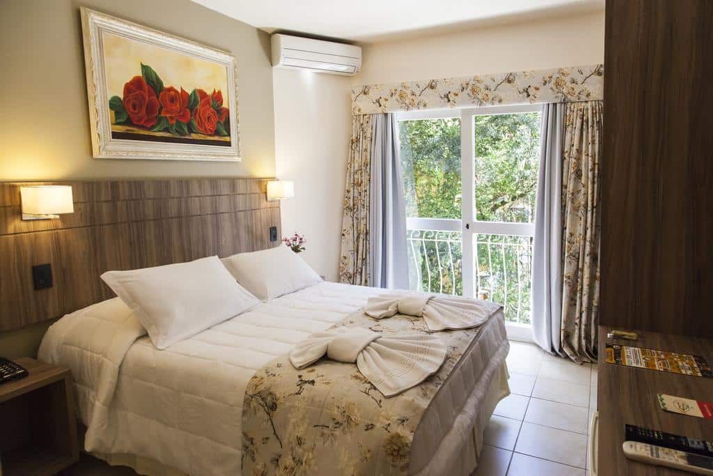 Suíte da Pousada Águia Branca com uma cama de casal à esquerda, um quadro de rosas pendurado acima da cabeceira e portas duplas com acesso a sacada privativa e vista para o jardim, para representar pousadas em Gramado.