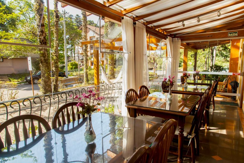 Refeitório da Pousada Águia Branca em Gramado com vista para o jardim e para rua. As paredes são envidraçadas e amplas, e do lado de dentro do refeitório há várias mesas retangulares com quatro cadeiras de madeira em cada.