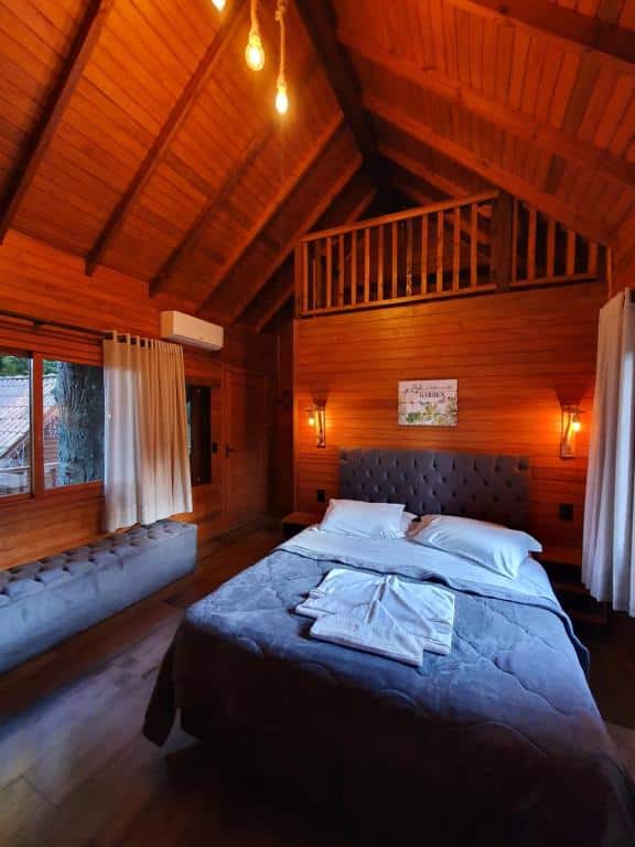 Suíte da Pousada Árvore da Coruja mostrando o lado interno de uma das cabanas com uma cama de casal e um pufe estofado cinza ao lado, para representar pousadas em Gramado.