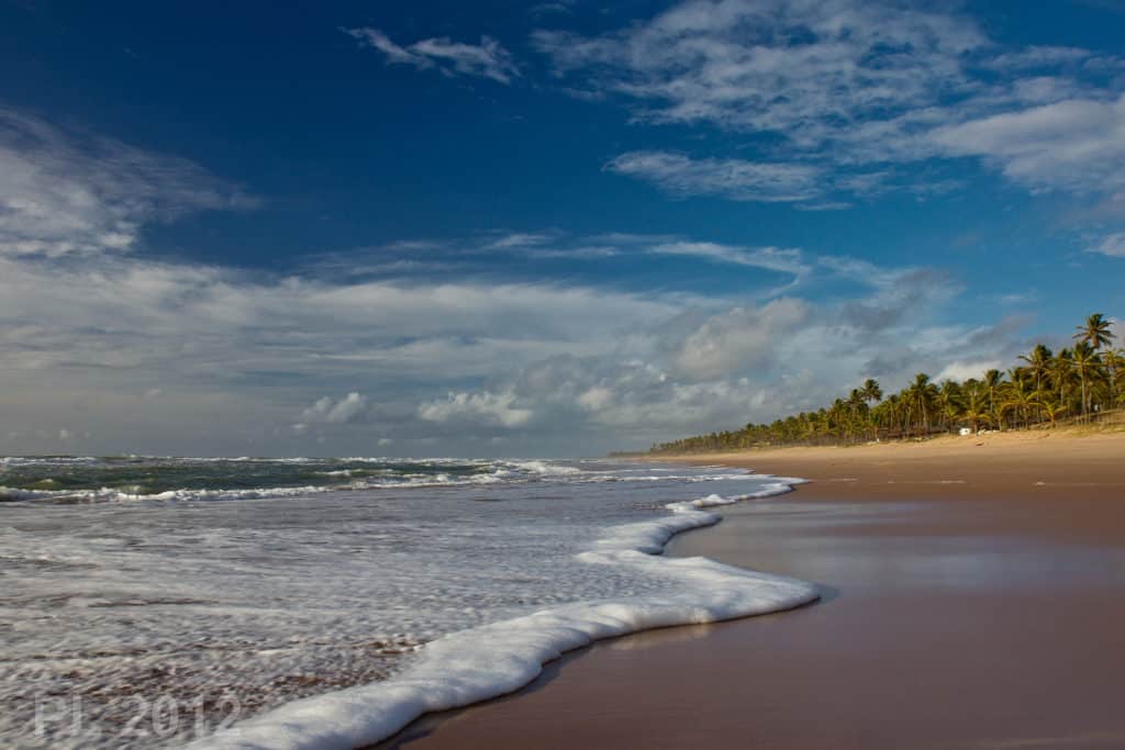 As ondas chegam a uma praia de areia com palmeiras sob um céu azul parcialmente nublado, na Praia de Imbassaí.