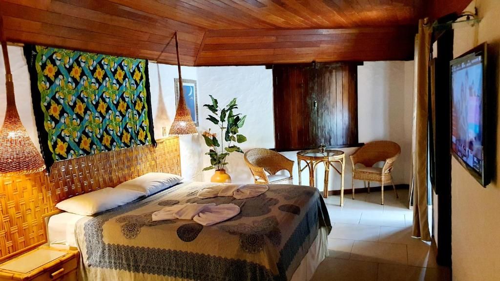 Um quarto aconchegante Pousada Entre as Águas com uma cama de casal, móveis de vime, uma TV na parede, iluminação decorativa e plantas, para representar pousadas em Imbassaí.