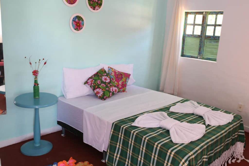 Uma cama bem arrumada com um cobertor xadrez verde, dois laços de toalha, travesseiros florais e uma mesa lateral redonda azul com um vaso, na Pousada Pelô de Imabassaí.
