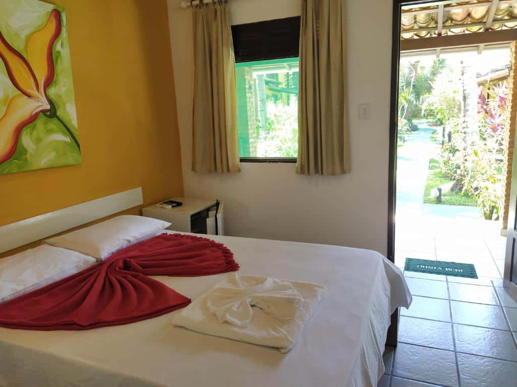 Quarto bem iluminado na Pousada Canto de Imbassaí com uma cama de casal, toalhas de arte, janela, porta aberta para um jardim ensolarado e piso de azulejos, para representar pousadas em Imbassaí.