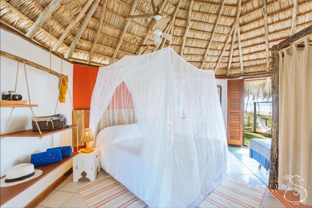 Cama com mosquiteiro em um quarto com telhado de palha, com porta aberta mostrando as palmeiras e a vista para o mar do lado de fora, na Pousada Capitú, para representar pousadas em Imbassaí.