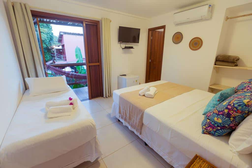 Quarto no Mar Ilha Pousada com uma cama de solteiro, uma cama de casal, TV, ar-condicionado, prateleiras abertas e uma porta que dá para uma varanda com rede, para representar pousadas em Itacaré.