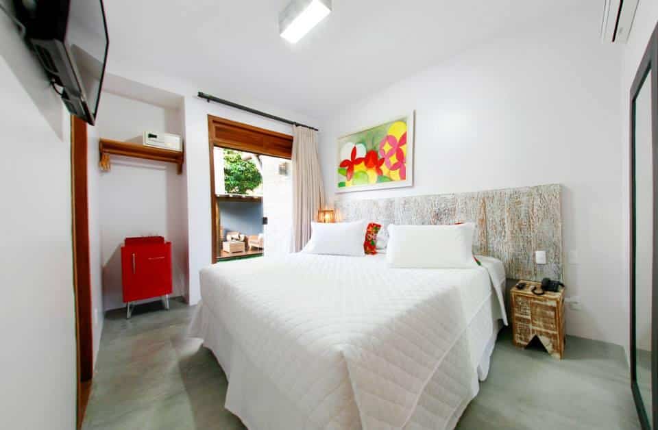 Uma cama bem arrumada em um quarto bem iluminado na Pousada Villa Maeva Itacaré com paredes brancas, uma pintura na parede e duas pequenas mesas laterais, para representar pousadas em Itacaré.