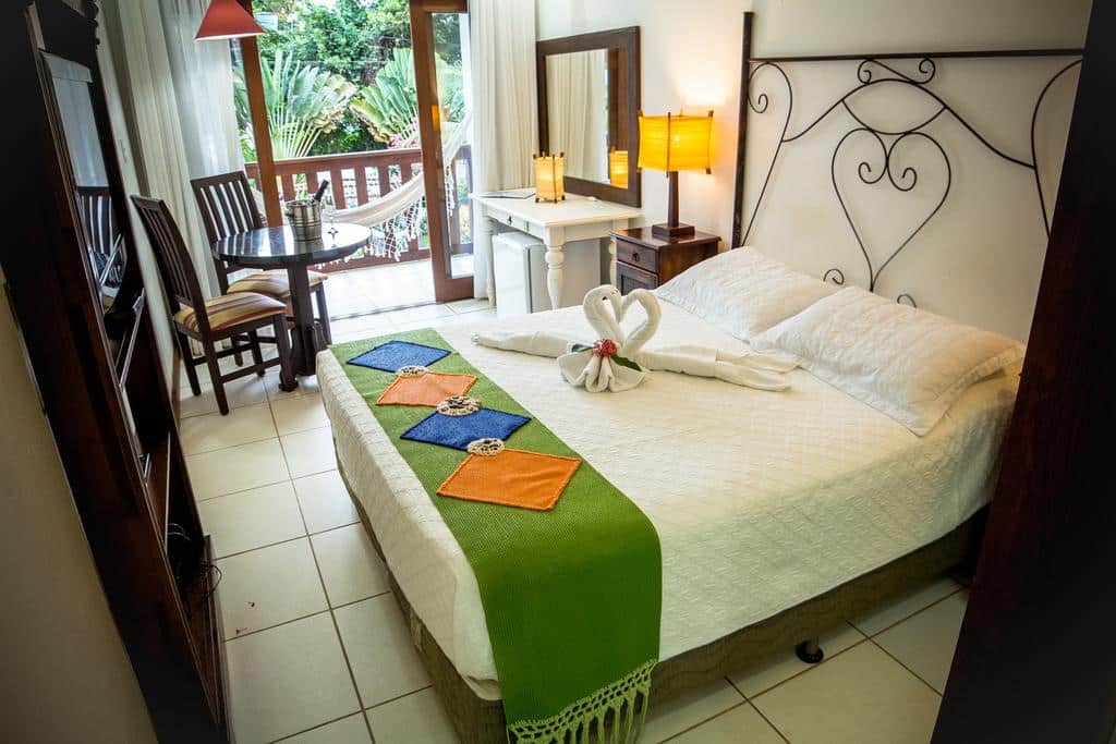 Um quarto no Porto dos Casais Guest House Itacaré com uma cama, toalha de cisne, mesa e cadeiras, luminárias e uma varanda com uma rede e vegetação externa, para representar pousadas em Itacaré.