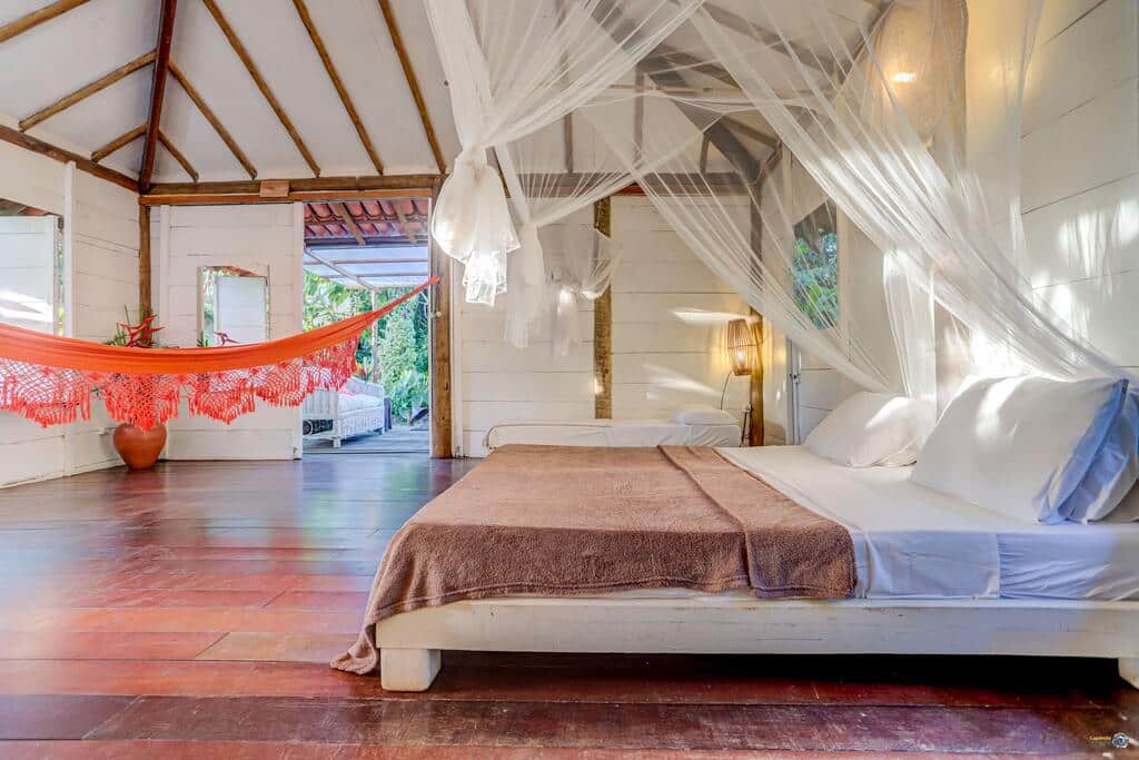 Um quarto espaçoso na Pousada Ilha Verde com uma cama grande sob um mosquiteiro branco e uma rede laranja perto de uma janela aberta, para representar pousadas em Itacaré.