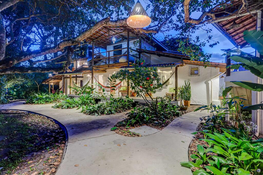 Casa de dois andares com janelas grandes, iluminação externa, telhado de telhas, cercada por plantas exuberantes e caminhos sinuosos de concreto ao anoitecer, na propriedade da Pousada Ilha Verde.