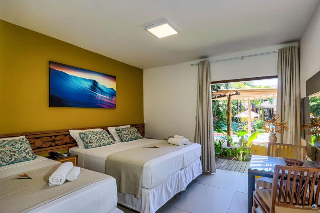 Um quarto na Pousada Vira Canoa com duas camas, uma escrivaninha e uma grande janela com cortinas com vista para um pátio com jardim, para representar pousadas em Itacaré.