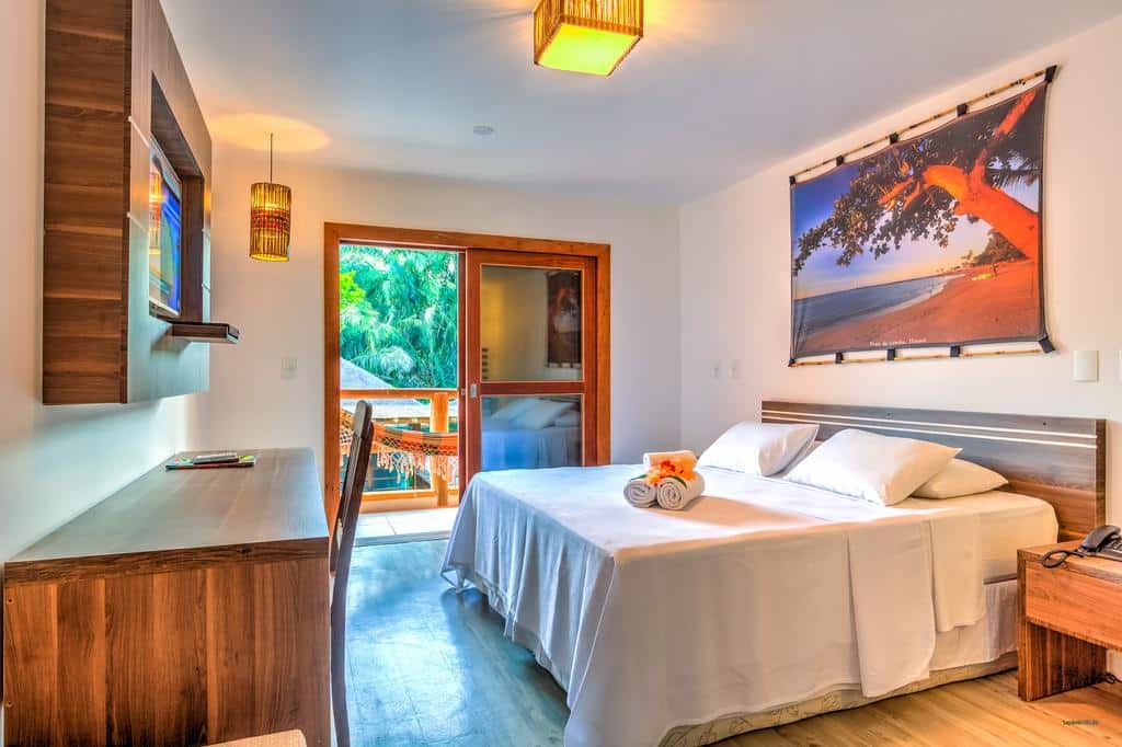 Quarto na Pousada Vila do Dengo bem iluminado com uma cama de casal, escrivaninha, arte na parede, TV e varanda com vista para as palmeiras, para representar pousadas em Itacaré.