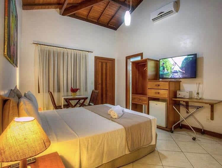 Uma cama bem arrumada em um quarto da Pousada Abaré com TV, móveis de madeira, frigobar e um abajur em uma mesa de cabeceira, para representar posuadas em Itacaré.