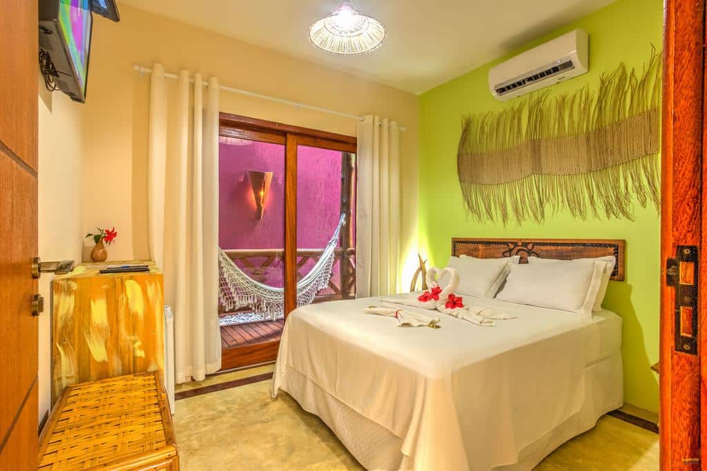 Quarto aconchegante no Raio de Sol Pousada com uma cama de casal, arte na parede, TV e portas de vidro deslizantes que levam a uma varanda com rede, para representar pousadas em Itacaré.