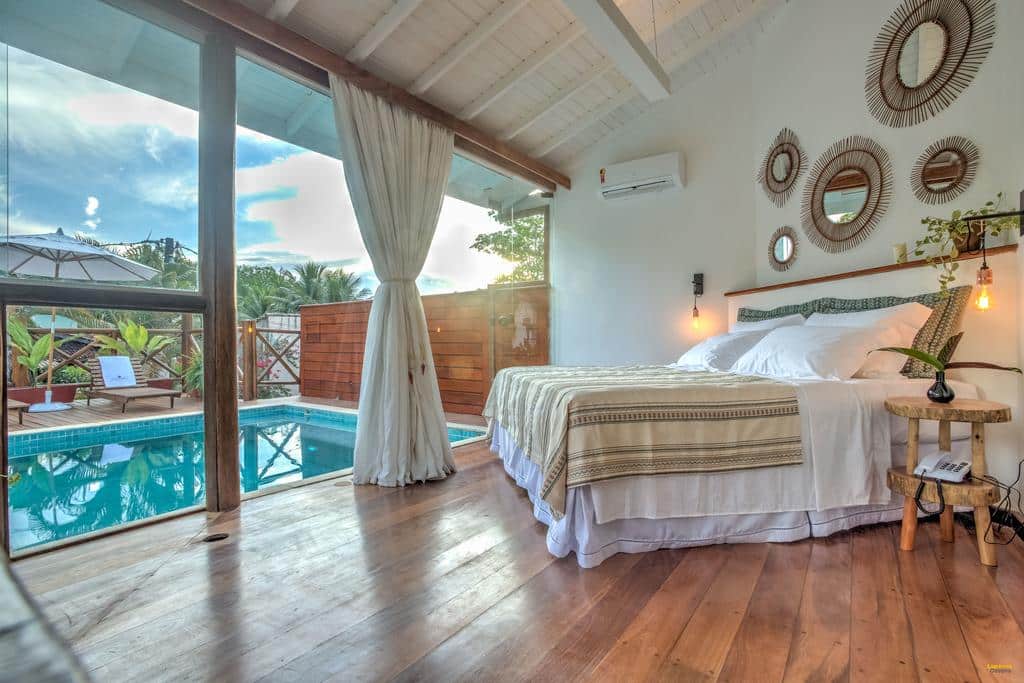 Um quarto em A Casa de Gabriella com uma cama ao lado da piscina, pisos de madeira, espelhos redondos na parede e janelas grandes com vista para o exterior, para representar pousadas em Itacaré.