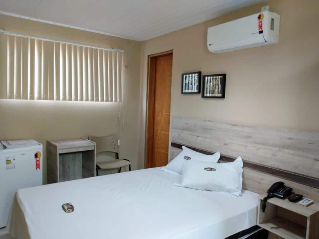 Uma cama bem arrumada em um quarto pequeno com frigobar, escrivaninha, cadeira, ar-condicionado e arte na parede acima da cama, na Pousada Café com Leitte, para representar pousadas em Juazeiro do Norte.