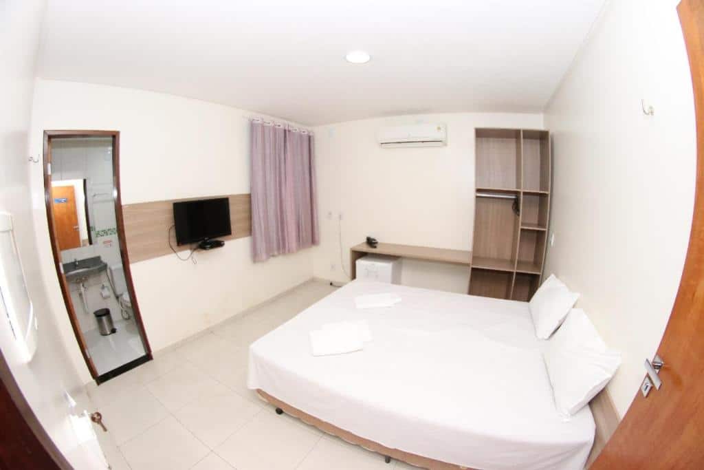 Um pequeno quarto na Pousada Santo André - O Apóstolo com uma cama, TV, armário aberto, frigobar e banheiro visível por uma porta aberta, para representar pousadas em Juazeiro do Norte.