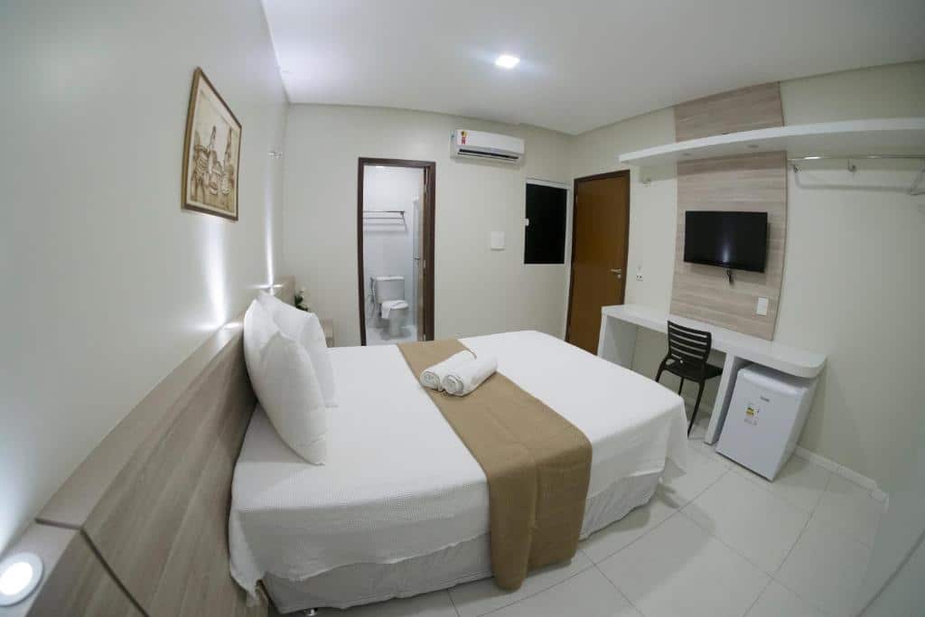 Um pequeno quarto do Hotel Padre Cícero com uma cama de casal, escrivaninha, frigobar, TV e um banheiro visível por uma porta aberta, para representar pousadas em Juazeiro do Norte.