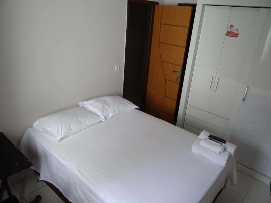 Uma cama bem arrumada com lençóis brancos, dois travesseiros e toalhas dobradas em um quarto simples com um guarda-roupa e uma porta aberta, na Pousada Vó Otília, para representar pousadas em Juazeiro do Norte.