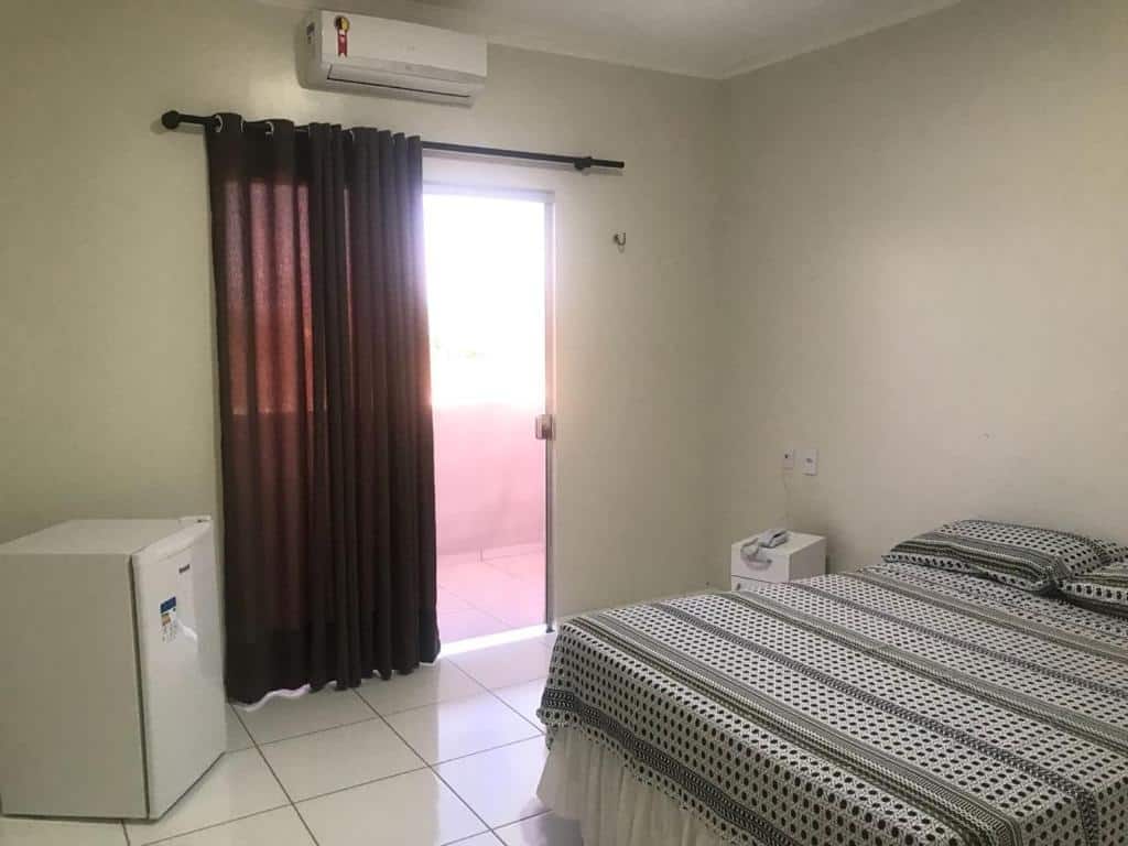 Um quarto simples na Pousada Aerogrill com uma cama, mesa de cabeceira, frigobar, ar-condicionado, cortinas escuras e uma porta de vidro para uma varanda, para representar pousadas em Juazeiro do Norte.