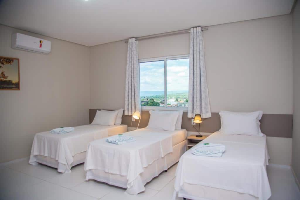 Três camas de solteiro com roupa de cama branca em um quarto limpo e iluminado com uma janela, cortinas e duas lâmpadas de cabeceira, no Hotel e Pousada Flor do Juá, para representar pousadas em Juazeiro do Norte.