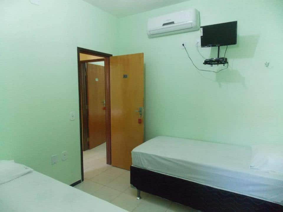 Um quarto simples com duas camas de solteiro, uma TV na parede e uma porta aberta que mostra um corredor com mais portas, na Pousada Cariri, para representar pousadas em Juazeiro do Norte.