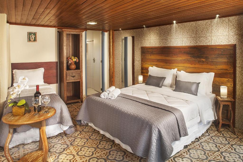 Quarto da Pousada Makutta, uma das pousadas em Lavras Novas, com uma cama de casal e uma cama de solteiro, teto de madeira, piso de azulejo, mesa de apoio com vinho e iluminação suave.