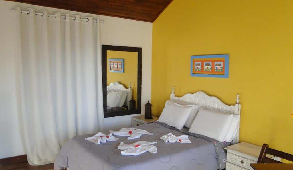 Uma cama bem arrumada da Pousada Vila do Campo, uma das pousadas em Lavras Novas, com cabeceira branca, parede amarela e um espelho que reflete parte do quarto.