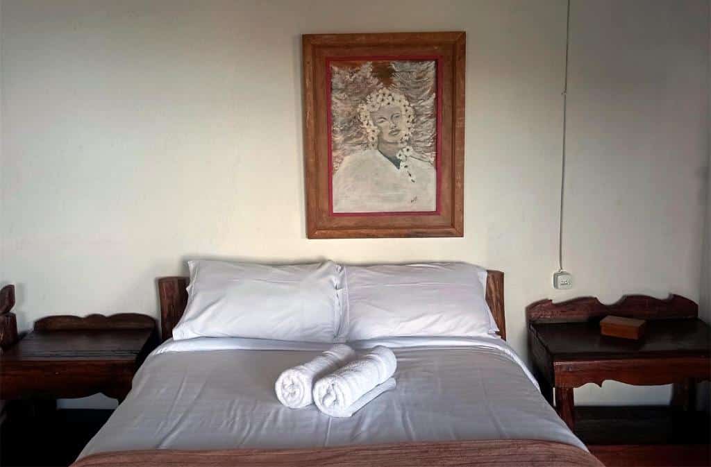 Uma cama bem arrumada com duas toalhas enroladas, mesinhas laterais de madeira e um retrato pendurado na parede acima da cabeceira. A imagem mostra a Pousada Solar dos Anjos, uma das pousadas em Lavras Novas