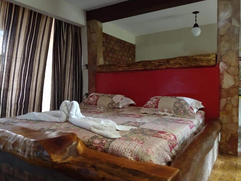 Quarto da Pousada Ecologica Vale das Cores, uma das pousadas em Lavras Novas, com uma cama de madeira rústica com roupa de cama de retalhos e cisnes de toalha, ao lado de cortinas listradas e uma luminária suspensa.