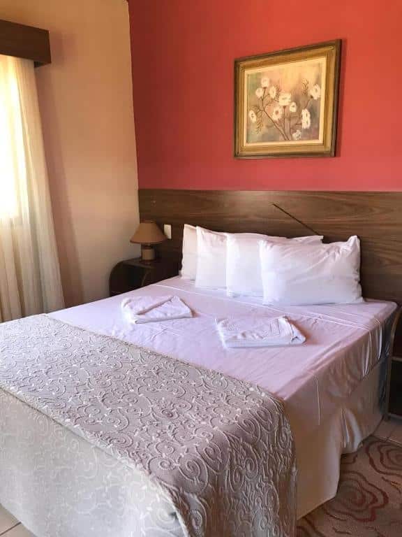Quarto da Caminho das Lavras, uma das pousadas em Lavras Novas, com uma cama de casal bem arrumada com lençóis brancos, duas toalhas e uma pintura floral em uma parede vermelha acima da cabeceira.