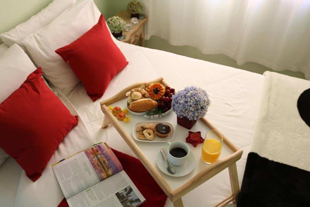 Uma bandeja de café da manhã com café, suco, pão e bolos fica sobre uma cama com travesseiros vermelhos e brancos e uma revista aberta. O local é a Biz & Biu Pousada Lavras Novas