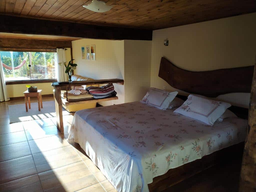Quarto da pousada Brisa de Lavras, em Lavras Novas, com uma cama de madeira, roupa de cama floral, um pequeno banco, um sofá e luz do sol que entra pelas grandes janelas.
