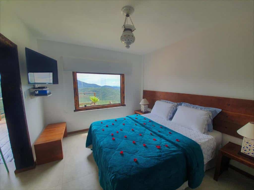 Uma cama de casal bem arrumada com roupa de cama azul e pétalas de rosas espalhadas em um quarto bem iluminado com janela e vista para a montanha. O local é a Mar De Montanhas, uma das pousadas em Lavras Novas