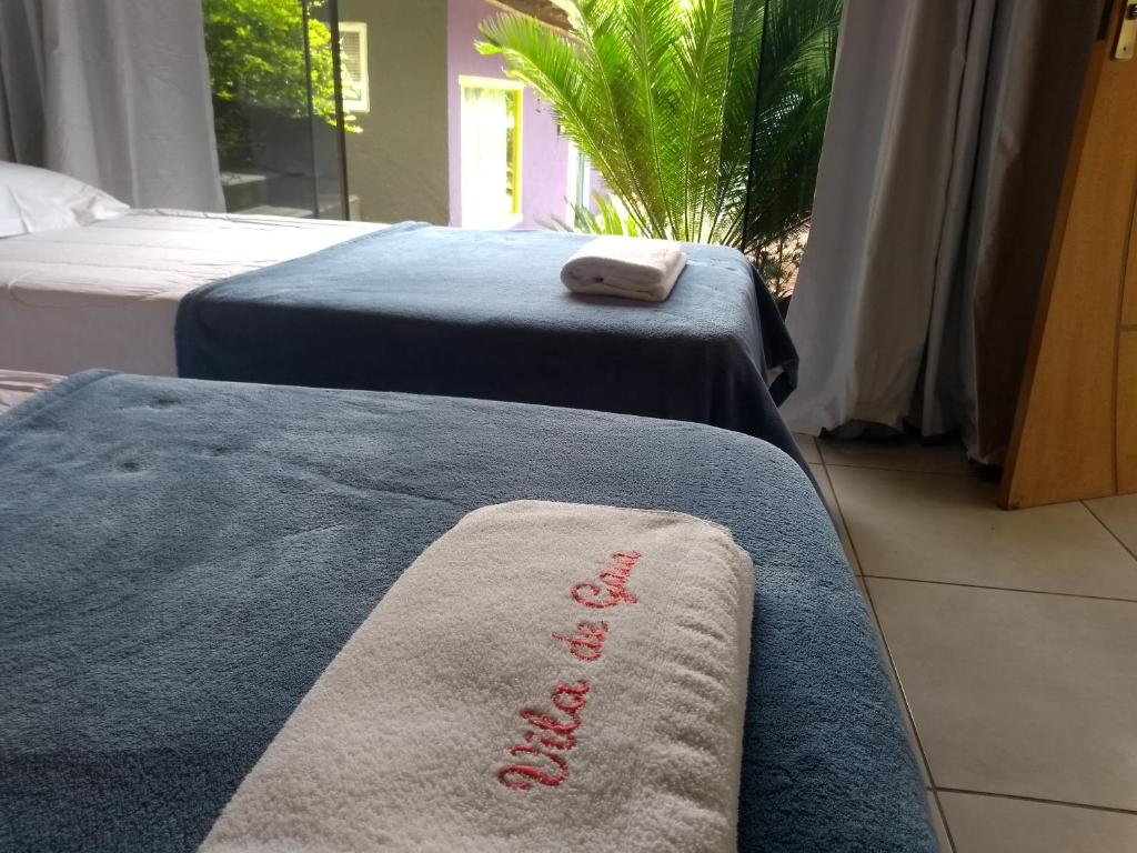 Duas camas com cobertores azuis e toalhas dobradas em um quarto bem iluminado com vista para a vegetação do lado de fora. O local é a Pousada Vila de Gaia, uma das pousadas em Lavras Novas