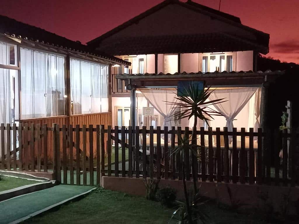 A imagem mostra a entrada da pousada Serra da Neblina em Lavras Novas, Minas Gerais, durante o entardecer. O céu está tingido de tons de rosa e roxo, indicando que o sol acabou de se pôr. A pousada possui uma construção rústica e charmosa, com detalhes em madeira. As janelas são amplas e estão fechadas com cortinas brancas, conferindo um ar acolhedor ao ambiente. A entrada da pousada é cercada por uma cerca de madeira baixa, e há um caminho de grama bem cuidado que leva até a porta principal. À direita, pode-se ver uma área externa coberta com um pergolado e cortinas, criando um espaço aconchegante para relaxar. Plantas ornamentais estão espalhadas pelo jardim, complementando a decoração natural do local. A iluminação interna da pousada está acesa, o que proporciona uma sensação de calor e hospitalidade.