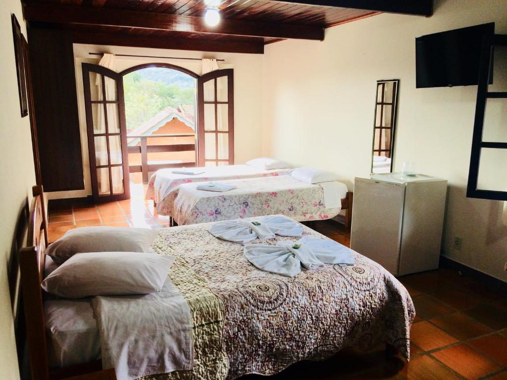 Quarto com duas camas, um pequeno frigobar, TV na parede, portas abertas para uma varanda e entrada de luz do dia, na Pousada Dona Ana, para representar pousadas em Monte Verde.