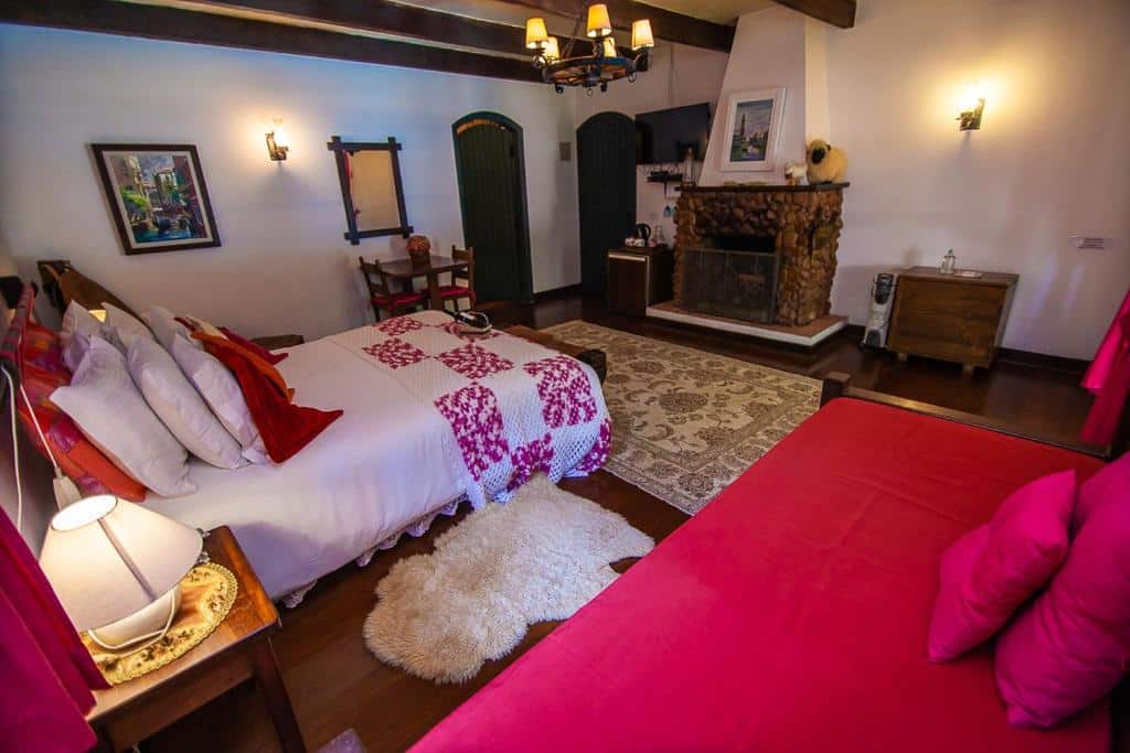 Um quarto aconchegante com duas camas, uma lareira de pedra, vigas de madeira e iluminação aconchegante; tapetes cobrem o piso de madeira, na Pousada Villa D'Amore, para representar pousadas em Monte Verde.