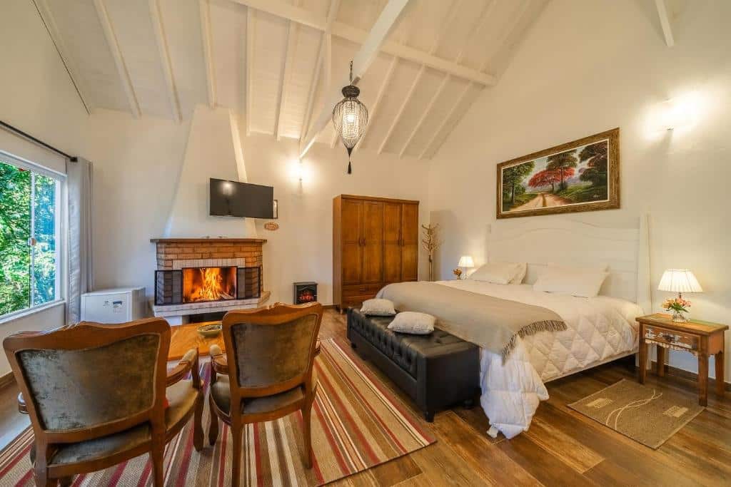 Quarto aconchegante da Pousada Jardim da Mantiqueira com lareira, cama, poltronas de madeira e TV. Um tapete colorido cobre o piso de madeira e quadros nas paredes, para representar pousadas em Monte Verde.